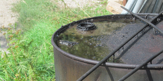 Wassergefährdende Stoffe Das Bild zeigt ein altes, rostiges Metallfass, das teilweise mit Wasser gefüllt ist. Auf der Wasseroberfläche liegen einige kleine Äste und Schmutz. Im Hintergrund sind grüne Pflanzen und ein heller Boden zu sehen