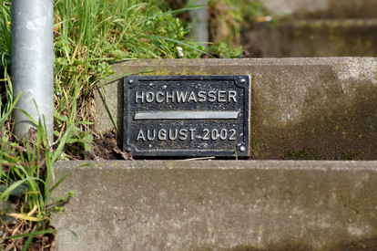 Risiko: Wie hoch ist unser Risiko? Kleine schwarze Metalltafel an einer Betontreppe. Aufschrift: „HOCHWASSER AUGUST 2002“. Leicht bewachsene Treppenumgebung.