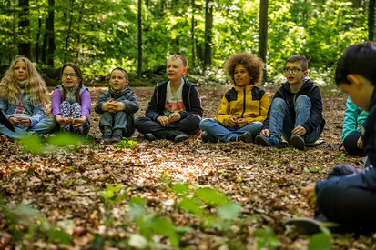 Wald – Lernen, Erleben, Erholen Kinder sitzen nebeneinander im Wald auf Laub