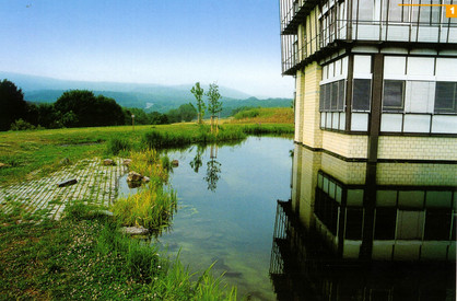 Umgang mit Niederschlagswasser Das Foto zeigt einen ruhigen kleinen Teich direkt neben einem modernen, mehrstöckigen Gebäude mit großen Fenstern und einer Fassade aus Glas und Metall. Das Gebäude spiegelt sich klar im Wasser des Teichs. Rund um den Teich wächst grünes Gras und einige Pflanzen. Im Hintergrund erstreckt sich eine weitläufige, grüne Landschaft mit sanften Hügeln und vereinzelten Bäumen.