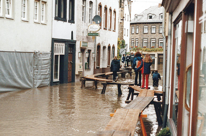 Vorsorge: Wie können wir uns vorbereiten? Überflutete Gasse in einer Altstadt. Holzbohlen als provisorischer Gehweg über dem Wasser. Mehrere Personen gehen auf den Bohlen. Alte Häuserzeilen links und rechts.