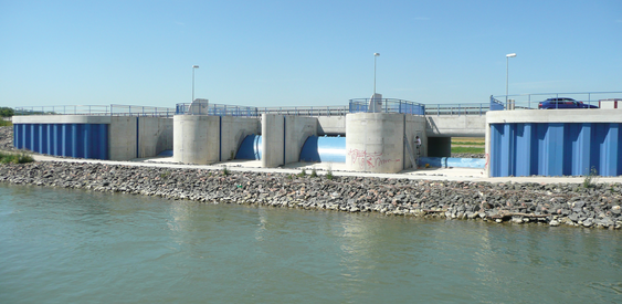 Management: Was macht das Land? Wasserbauanlage mit Betonstruktur und blauen Metallteilen. Wasser fließt durch Öffnungen im Bauwerk. Kiesufer im Vordergrund, klarer Himmel im Hintergrund.