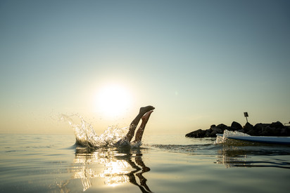 Öffentlichkeitskampagne der Wasserwirtschaft Person springt kopfüber ins Wasser. Nur die Beine ragen aus dem Wasser. Die Sonne steht tief am Himmel und spiegelt sich auf der Wasseroberfläche. Rechts im Bild sind Felsen und ein Paddleboard zu sehen.