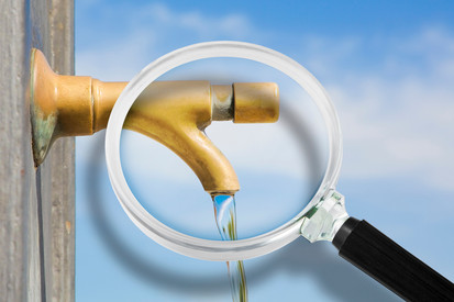 Trinkwasser Überwachung Das Bild zeigt einen goldfarbenen Wasserhahn, an dem Wasser fließt. Ein Vergrößerungsglas ist so positioniert, dass es den Wasserhahn und den Wasserstrahl vergrößert darstellt. Der Hintergrund ist ein blauer Himmel mit wenigen Wolken.