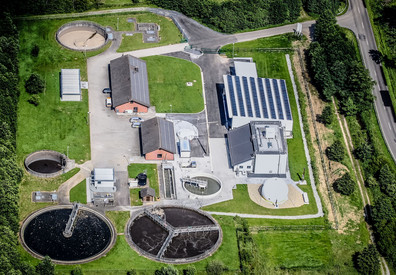 Kommunales Abwasser Das Bild zeigt eine Luftaufnahme einer Kläranlage mit mehreren runden Becken und Gebäuden. Die runden Becken sind unterschiedlich groß und enthalten jeweils Wasser oder Schlamm. Die Gebäude, meist mit dunklen Dächern und einigen mit Solarpaneelen, stehen auf gepflasterten Flächen zwischen den Becken. Die gesamte Anlage ist von grünen Rasenflächen und Hecken umgeben.