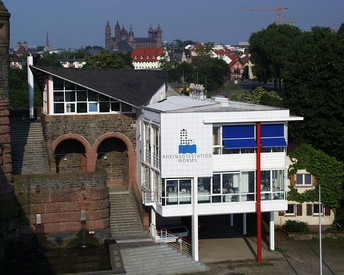 Gewässerschutz Weißes modernes Gebäude mit großen Fenstern und roten Säulen – zeigt die Rheingütestation Worms. Steht neben alter Backsteinmauer mit Bögen. Im Hintergrund Bäume und historische Gebäude.