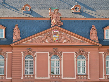 Historisches Gebäude mit rosafarbenem Fassadenschmuck. Drei große Fenster mit weißen Rahmen. Oben Relief mit Figuren und Verzierungen, mittig Statue eines sitzenden Mannes. Blauer Himmel als Hintergrund.